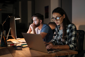 Two business professionals working late on laptops and computers in an office